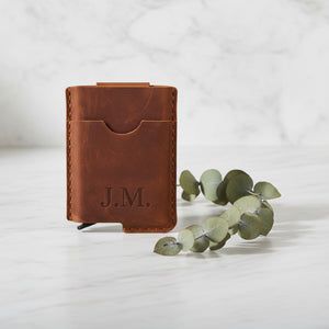 Brown leather card holder engraved with J.M. initials on marble with eucalyptus