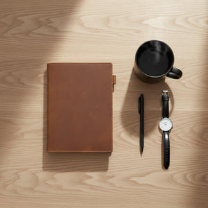 Brown leather journal cover styled with a pen, watch and coffee on a wood desk